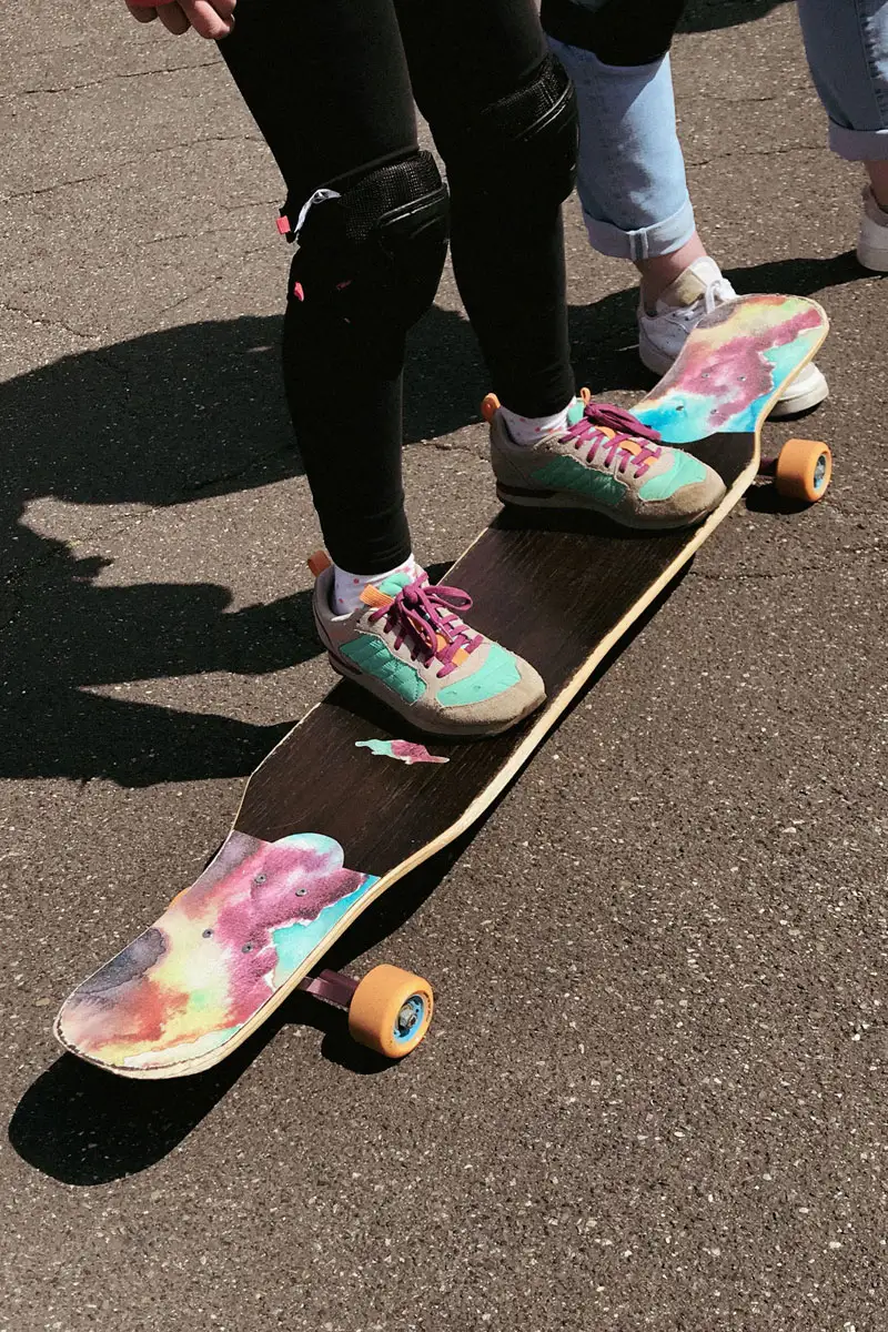 Ein Longboard mit farbigem Griptape, auf dem ein Kind steht. Man sieht nur die Füße des Kindes