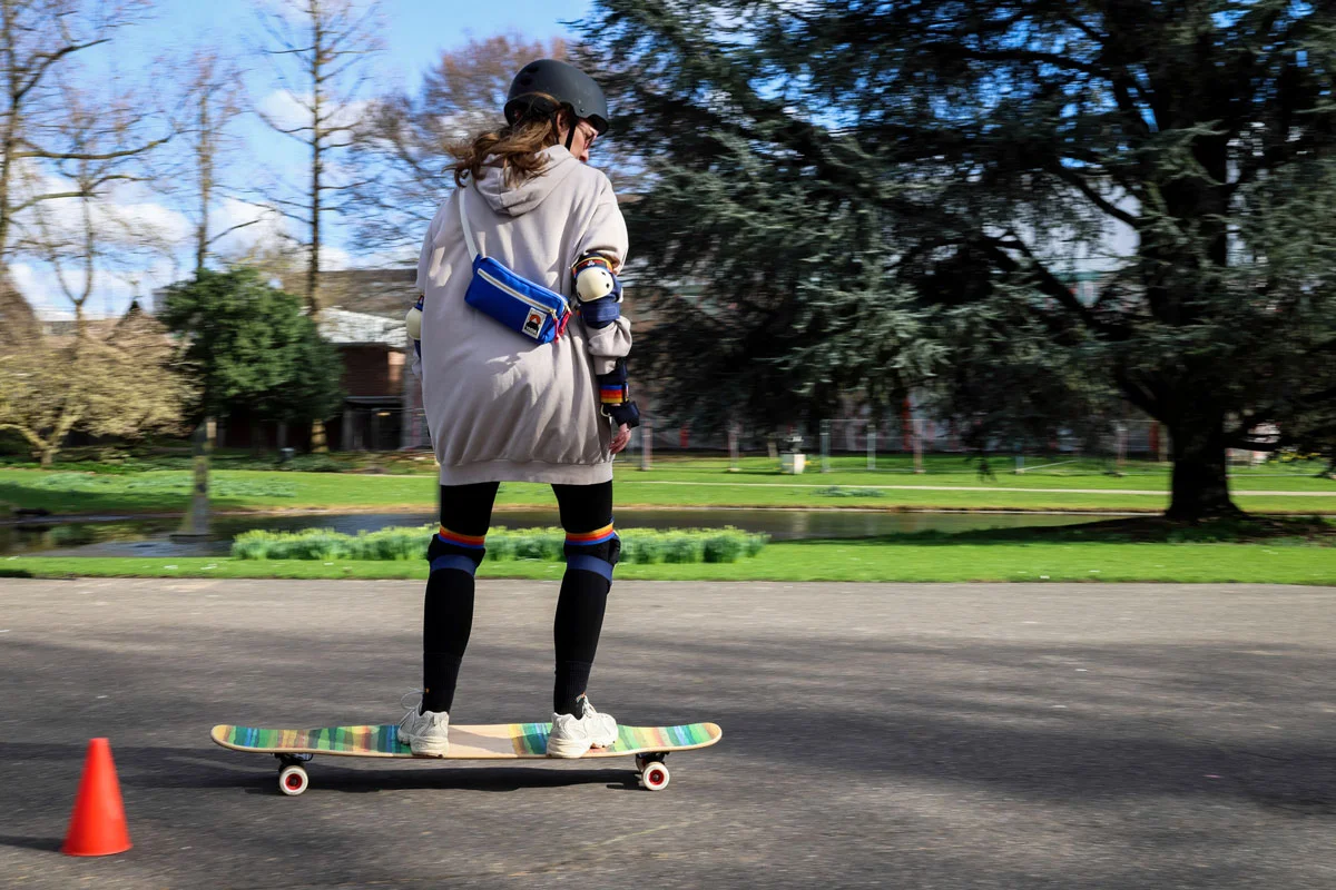 Eine Frau steht auf einem Longboard beim Longboard Kurs in Köln. Links von ihr ist ein orangefarbenes Hütchen zu sehen, im Hintergrund ein Weiher und Wiese